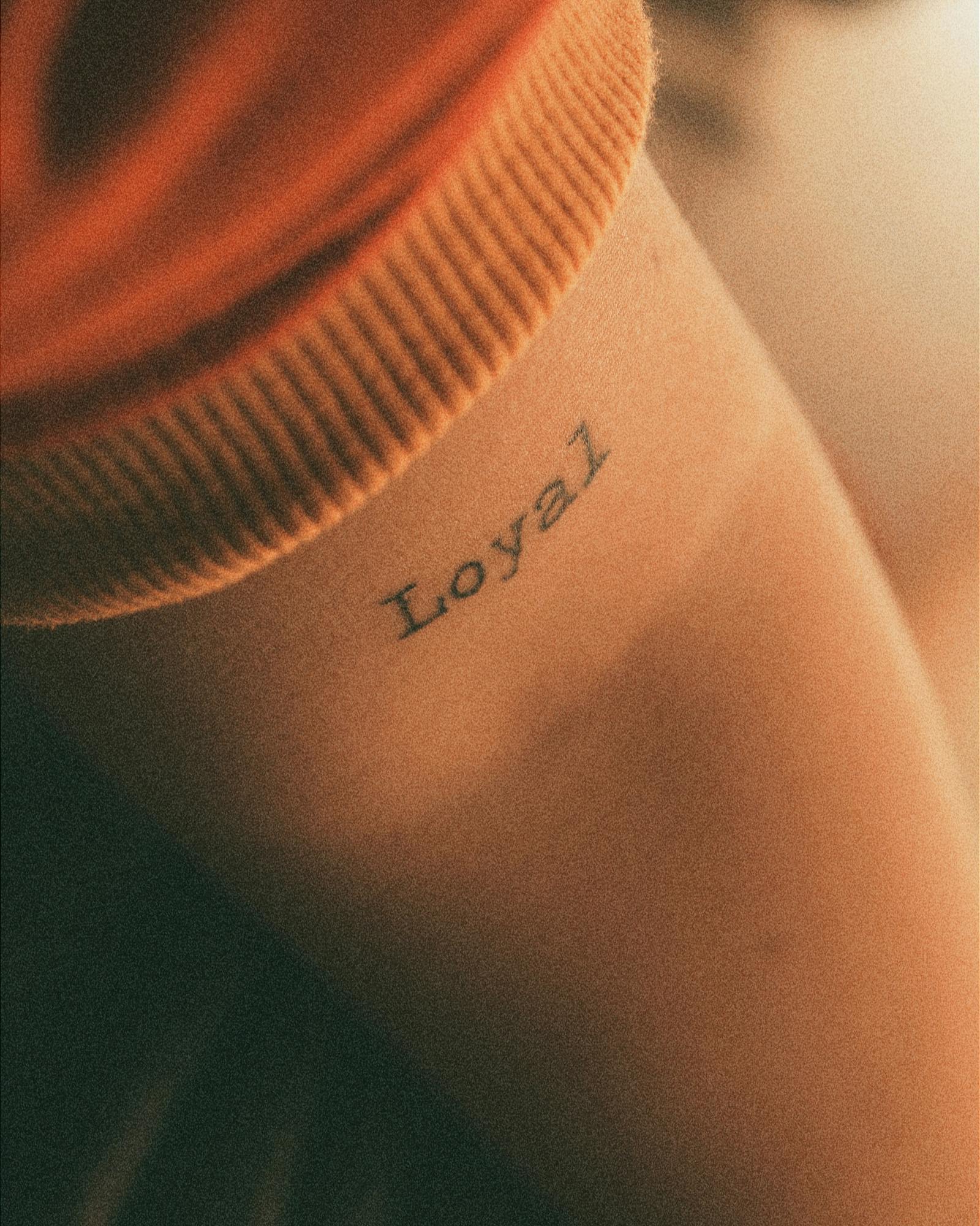 Hand-lettered script tattoo reading 'Loyal' on a forearm, warm tones